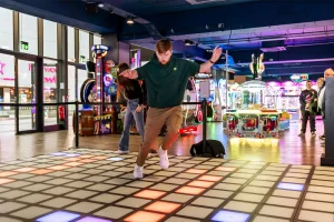 A wide-angle view of a futuristic immersive game room featuring an interactive LED floor grid for family entertainment centers.
